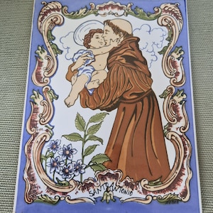 Op de afbeelding: Een rechthoekige keramische wandplaat met een kleurrijke illustratie van de heilige Antonius die het kindje Jezus vasthoudt. De scène is omlijst door een sierlijke rand met blauwe accenten en bevat onderaan de tekst SANTO ANTONIO.