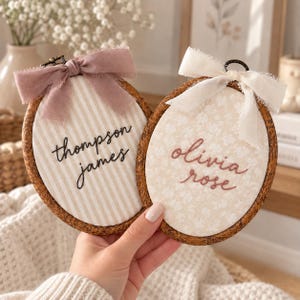 Puede incluir: Dos bastidores de bordado ovalados con marcos de madera oscura. El bastidor izquierdo presenta rayas beige, el texto bordado en negro 'thompson james' y un lazo malva. El derecho tiene tela floral crema, 'olivia rose' en texto rosa y un lazo blanco.
