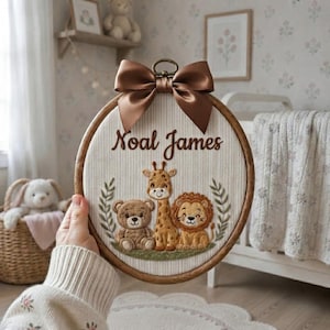 Puede incluir: Un bastidor de bordado circular de madera con un fondo de tela color crema y el nombre Noal James bordado en letra cursiva marrón. Debajo del nombre hay figuras cosidas de un oso pardo, una jirafa y un león, rematadas con un lazo de satén marrón.