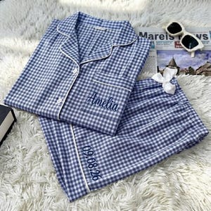 May include: A blue and white gingham pajama set featuring a button-down shirt with a collar and a chest pocket embroidered with the name Amelia. The matching shorts include a white ribbon bow and the embroidered date 09.08.26 on the hem.