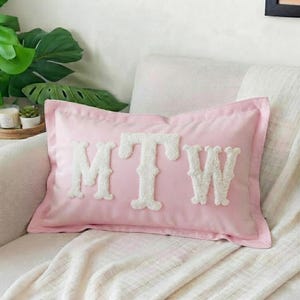 Könnte beinhalten: Ein rechteckiges, hellrosa Dekokissen mit den weißen Buchstaben MTW in einer strukturierten, flauschigen Chenille-Applikation, die mittig auf der Vorderseite angebracht sind. Das Kissen hat einen schlichten, abgesteppten Rand und eine glatte Oberfläche.