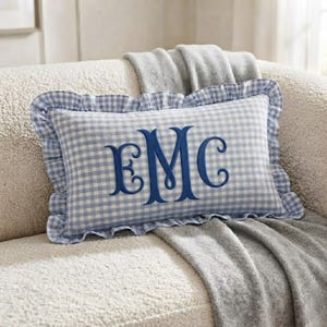 May include: A rectangular throw pillow featuring a light blue and white gingham pattern with a ruffled edge. The center of the pillow displays a large, dark blue embroidered monogram with the letters E, M, and C in a decorative script font.