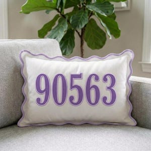 Könnte beinhalten: Ein rechteckiges weißes Zierkissen mit den mittig aufgestickten violetten Zahlen 90563. Das Kissen besitzt einen dekorativen Wellenrand, der mit einer passenden violetten Paspelierung versehen ist und einen deutlichen Rahmen um das Kissen bildet.
