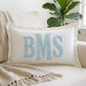 May include: A rectangular white throw pillow featuring the letters BMS in large, light blue, textured, tufted embroidery centered on the front. The pillow has a clean, simple border design and is made of a smooth, solid-colored fabric material.