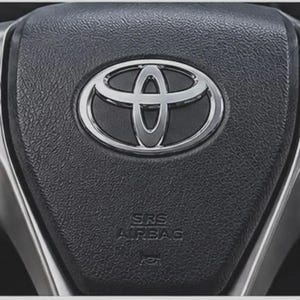 May include: A black textured steering wheel center featuring a prominent chrome Toyota logo emblem. The assembly includes silver-toned side panels with integrated control buttons labeled with symbols, MODE, HOLD, and DISP, plus an SRS AIRBAG label.