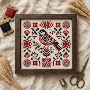 Op de afbeelding: Een vierkant kruissteek borduurwerk in een donkere houten lijst met een vogel op een tak, omringd door symmetrische rode en zwarte bloem- en geometrische patronen op een crèmekleurige stoffen achtergrond, getoond met garenklosjes.