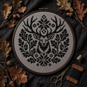 May include: A circular wooden embroidery hoop containing a cross-stitch pattern on grey fabric. The design features a black stag head centered among oak leaves, acorns, and geometric snowflake motifs, creating a symmetrical nature-inspired image.