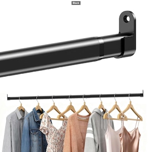 May include: A matte black adjustable tension rod featuring a cylindrical design with a mounting bracket at the end. The rod is shown installed horizontally, supporting several wooden hangers with various garments including shirts, a jacket, and dresses.