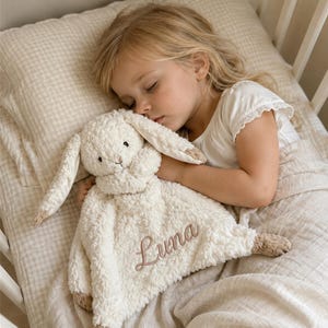 Puede incluir: Una manta de seguridad de felpa color crema con una cabeza de conejo de peluche de orejas largas y el nombre Luna bordado en marrón. La tela suave y texturizada incluye pequeños detalles en marrón en las orejas y en las esquinas de la manta.