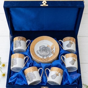 May include: A set of six white porcelain coffee cups and saucers featuring a gray rose motif and gold trim, presented in a deep blue velvet-lined display box with a gold latch. The cups and saucers are arranged neatly within the blue fabric interior.