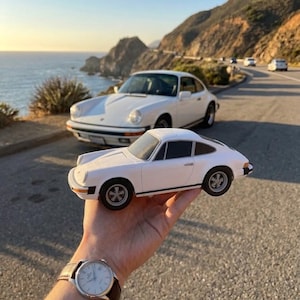 Può includere: Un modellino di auto bianco con una classica silhouette coupé, finiture dei finestrini nere e ruote grigio scuro con un centro a forma di stella. Il modello presenta una striscia laterale nera e indicatori di direzione anteriori arancioni.