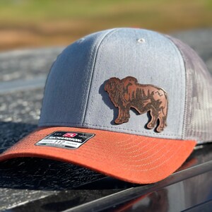 May include: A gray and black trucker hat with a brown leather buffalo patch. The hat has a red brim and a mesh back.