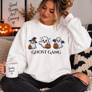 Könnte beinhalten: Ein weißes Langarm-Sweatshirt mit einer Grafik von drei Cartoon-Geistern mit Kürbissen und dem Text GHOST GANG. Der mittlere Geist wird von einem Geist mit Hexenhut und einem Geist mit Fledermausflügeln flankiert. Eine weiße Tasse mit Spooky Season Text.