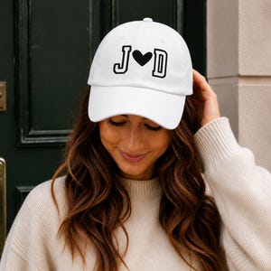 Könnte beinhalten: Eine weiße Baseballkappe mit den schwarz gestickten Buchstaben J und D, die durch ein schwarzes Herzsymbol in einer Schriftart im College-Stil getrennt sind. Die Kappe hat einen gebogenen Schirm und eine strukturierte Krone mit Belüftungsösen.