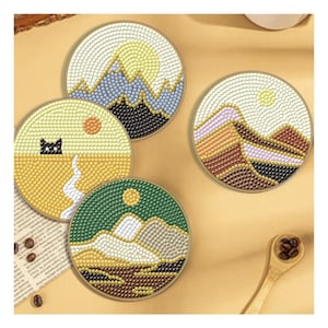 Puede incluir: Cuatro posavasos circulares con diseños de paisajes en diamond painting. Uno muestra una montaña con un sol amarillo, otro un gato en un desierto amarillo, un tercero con montañas marrones y el cuarto un paisaje verde con una cima blanca.