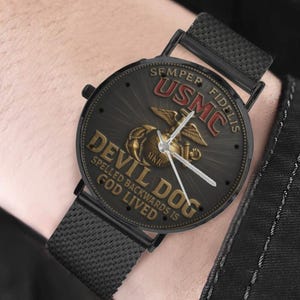 Könnte beinhalten: Eine schwarze Armbanduhr mit Mesh-Armband und rundem Zifferblatt, das das USMC-Emblem mit Adler, Weltkugel und Anker zeigt. Das Zifferblatt enthält den Text USMC, SEMPER FIDELIS, DEVIL DOG und SPELL ED BACKWARDS IS GOD LIVED in Gold und Rot.