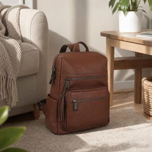 May include: A medium brown leather backpack featuring a textured surface, two front zippered compartments, and a decorative tassel attached to the side zipper. The bag includes dark metal hardware, adjustable shoulder straps, and side pockets.