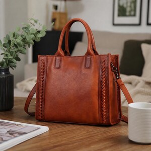 May include: A structured cognac-coloured leather handbag featuring two top handles and a detachable shoulder strap. The bag has a smooth central panel flanked by vertical braided leather detailing on both sides, with dark metal hardware attachments.