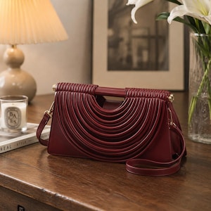 May include: A deep burgundy handbag featuring a structured rectangular base and a distinctive front panel adorned with multiple concentric, raised, semi-circular ridges. The bag includes a top handle and a thin, matching shoulder strap with gold hardware.