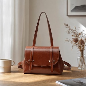 Könnte beinhalten: Eine strukturierte braune Ledertasche mit abgerundetem Klappverschluss, zwei vertikalen Zierriemen mit silberfarbenen Metallnieten und zwei hohen Schulterriemen. Die Tasche hat eine glatte, polierte Oberfläche und ein klassisches rechteckiges Design.