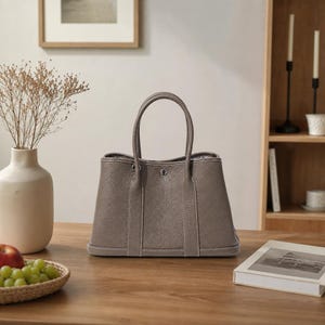 May include: A taupe-coloured tote bag with a textured leather-like finish, featuring two vertical straps, white contrast stitching, and a central silver-toned metal snap closure. The bag has a structured rectangular base and two top handles.