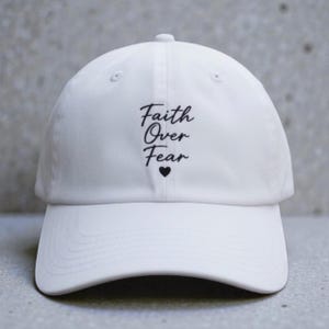 Könnte beinhalten: Eine weiße Baseballkappe mit dem schwarz gestickten Text Faith Over Fear in einer kursiven Schriftart, platziert über einem kleinen schwarzen Herzsymbol in der Mitte der Vorderseite der Kappe, die einen gebogenen Schirm und Ösen aufweist.