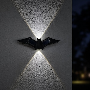 May include: A black bat-shaped wall light featuring integrated solar panels on the wings. The fixture emits two distinct beams of bright white light, one directed upward and one directed downward, creating a symmetrical pattern on the wall surface.