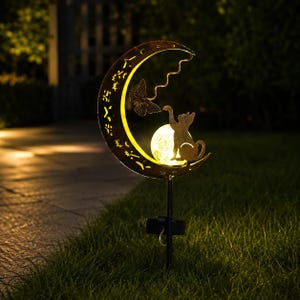 May include: A solar-powered garden stake featuring a crescent moon silhouette with cut-out star patterns. A metal cat figure reaches toward a glowing, textured glass orb light positioned within the moon, while a butterfly rests nearby.