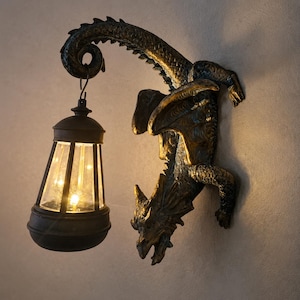 Può includere: Una scultura da parete a forma di drago con finitura bronzo scuro e riflessi dorati, dotata di una coda curva che sostiene una lanterna appesa. La lanterna presenta una cornice nera con pannelli in vetro trasparente e una luce calda.