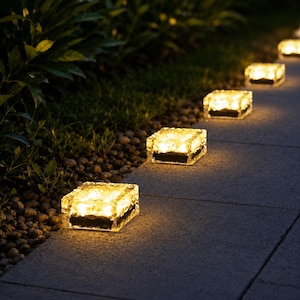 May include: A series of square, clear glass solar lights arranged in a line along a stone pathway. Each light features a textured, ice-like surface that emits a warm, glowing yellow light, illuminating the ground and the adjacent garden area.