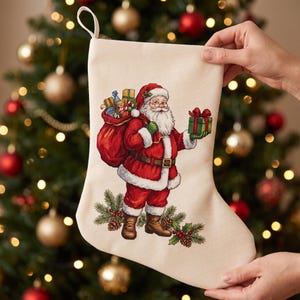 Op de afbeelding: Een crèmekleurige kerstsok van stof met een kruissteekontwerp van de Kerstman in een rood pak met witte randen, die een grote rode zak vol kleurrijke cadeaus draagt en een klein groen cadeau doosje met een rode strik vasthoudt.