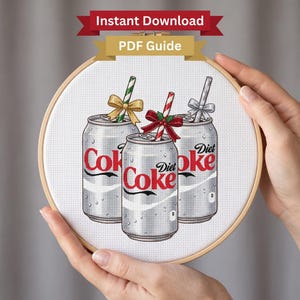 Könnte beinhalten: Ein Kreuzstichmuster in einem hölzernen Stickrahmen mit drei silbernen Diet Coke Dosen. Jede Dose hat einen Strohhalm mit einer dekorativen Schleife: eine goldene Schleife mit grünem Strohhalm, eine rote Schleife mit rot-weißem Strohhalm und eine silberne Schleife mit silbernem Strohhalm.