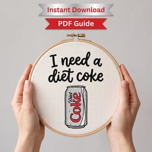 以下が含まれることがあります： 木製の円形刺繍枠に白い布が張られ、黒い刺繍で「I need a diet coke」という文字が施されています。その下には、赤と白の文字が書かれた銀色のダイエットコークの缶のグラフィックが布の中央に配置されています。