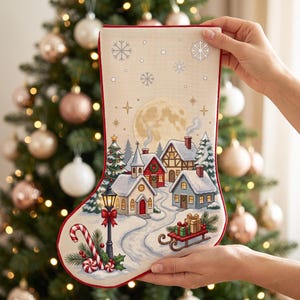 Peut inclure: Une chaussette de Noël couleur crème ornée d'un motif de village hivernal imprimé avec des maisons enneigées, une église, un lampadaire, un traîneau avec des cadeaux et des sucres d'orge. Le design inclut une grande lune et des flocons.