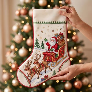 Puede incluir: Un calcetín navideño de color crema con un diseño de punto de cruz que muestra a Santa Claus en un trineo rojo tirado por dos renos. La escena incluye pinos verdes, copos de nieve y un saco de regalos, todo bordeado con ribete rojo.