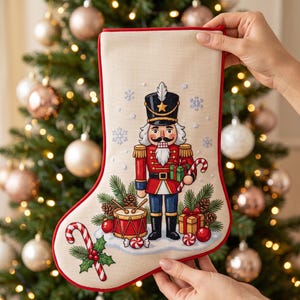 May include: A cream-coloured Christmas stocking featuring a cross-stitch design of a nutcracker soldier, a drum, a gift box, pine branches, pinecones, red ornaments, and candy canes, all bordered by a vibrant red trim against a neutral fabric.