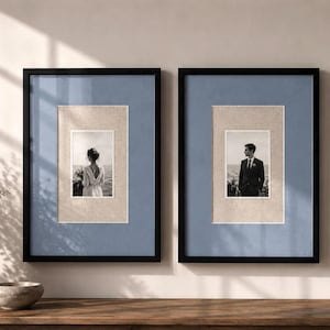 May include: Two rectangular black picture frames featuring blue matting and beige inner borders. Each frame contains a black and white photograph, one showing a person in a dress from behind and the other showing a person in a suit.