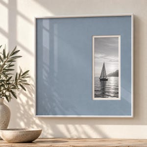 Op de afbeelding: Een vierkante witte fotolijst met een brede, stoffig blauwe passe-partout. Binnenin bevindt zich een verticale rechthoekige zwart-witfoto van een zeilboot op kalm water bij zonsondergang, met bergen zichtbaar op de verre achtergrond.