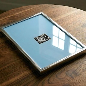 May include: A rectangular picture frame with a metallic silver finish and a light blue mat board. The frame contains a small, centered black and white photograph depicting two people sitting at a table, protected by a clear glass front panel.