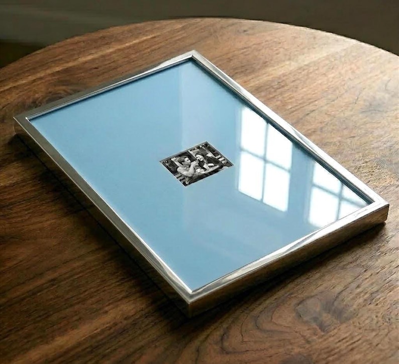 May include: A rectangular picture frame with a metallic silver finish and a light blue mat board. The frame contains a small, centered black and white photograph depicting two people sitting at a table, protected by a clear glass front panel.