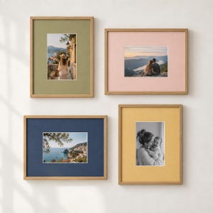 Op de afbeelding: Een set van vier houten fotolijsten met gekleurde passe-partouts. De lijst linksboven is olijfgroen met een kustscène, rechtsboven is lichtroze met een berglandschap, linksonder is marineblauw met zeezicht en rechtsonder is mosterdgeel met een zwart-witportret van een persoon die een baby vasthoudt.