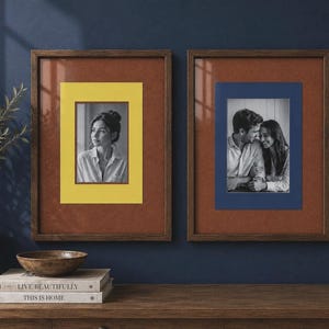 May include: Two rectangular dark wood picture frames with glass fronts. The left frame features a yellow mat surrounding a black and white portrait of a person. The right frame has a blue mat surrounding a black and white photo of a couple.