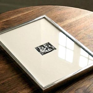 May include: A rectangular picture frame with a thin, polished silver-toned metal border. The frame contains a large, off-white mat board with a small, centered black and white photograph depicting two people sitting at a table with drinks.