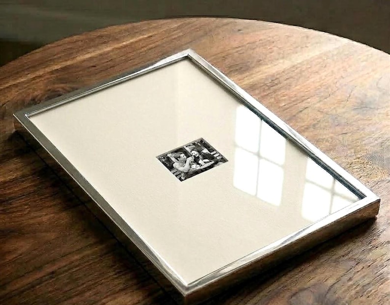 May include: A rectangular picture frame with a thin, polished silver-toned metal border. The frame contains a large, off-white mat board with a small, centered black and white photograph depicting two people sitting at a table with drinks.