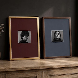 May include: Two rectangular picture frames with thin borders, one gold and one dark wood, each containing a centered black and white portrait photograph. The gold frame features a deep burgundy mat, while the dark wood frame has a slate blue mat.