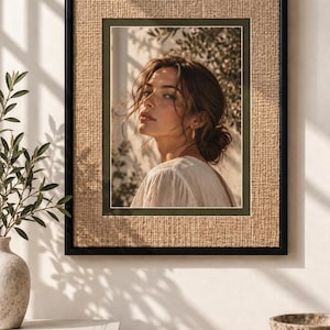 Op de afbeelding: Een rechthoekig wandkunstwerk met een portret van een persoon met donker haar in een losse knot, gekleed in een wit bovenstuk. Het portret is omgeven door een donkergroene passe-partout en een geweven beige rand, gevat in een dunne zwarte lijst.