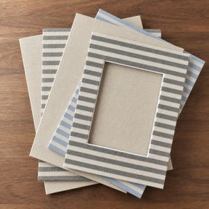 Puede incluir: Una pila de marcos de fotos rectangulares que presentan una mezcla de cubiertas de tela beige sólida y cubiertas de tela con rayas horizontales en tonos gris y azul claro. Cada marco tiene un recorte rectangular central para fotos.