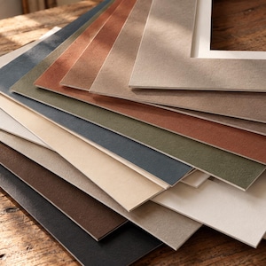 Puede incluir: Una colección de paspartús rectangulares para marcos de fotos dispuestos en forma de abanico. Los paspartús presentan una variedad de colores sólidos que incluyen negro, marrón oscuro, crema, beige, verde oliva, azul marino, terracota y gris claro.