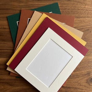 May include: A stack of rectangular paper picture mats in various colors including dark green, brown, tan, yellow, deep red, and off-white. Each mat features a centered rectangular cutout with a beveled edge, designed for framing artwork or photos.