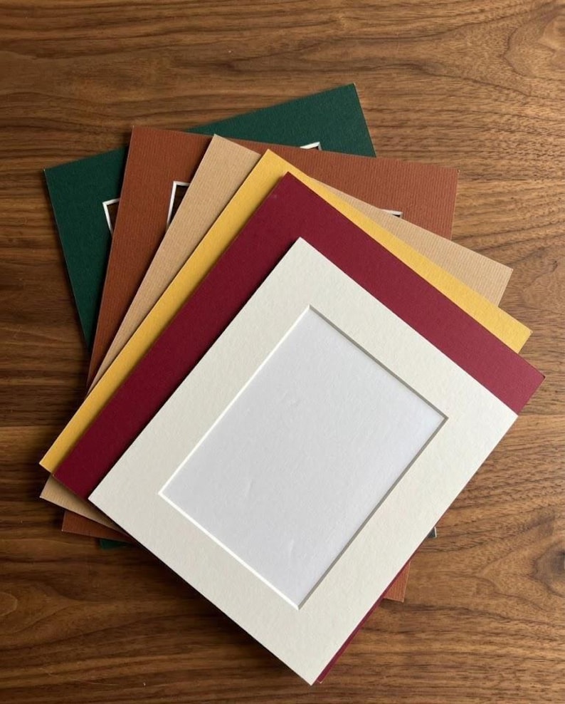 May include: A stack of rectangular paper picture mats in various colors including dark green, brown, tan, yellow, deep red, and off-white. Each mat features a centered rectangular cutout with a beveled edge, designed for framing artwork or photos.